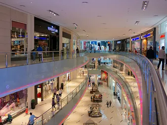  An interior, multi-level view of a modern, brightly lit shopping mall with a curved, open design. The mall features several floors of retail stores with glass railings around the balconies. People are visible walking on all levels, browsing the shops, and visiting small kiosks in the centre of the ground floor. 